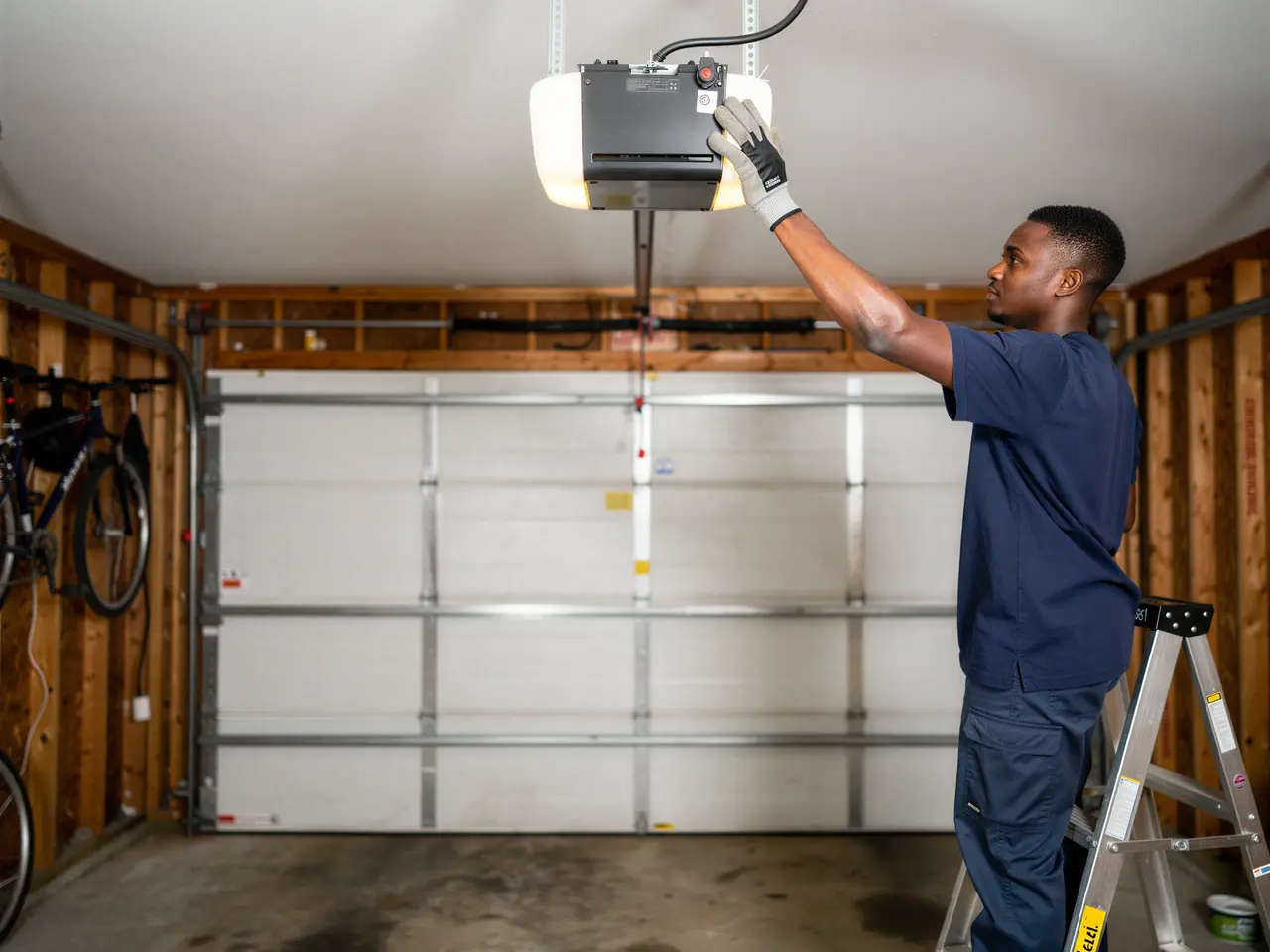 A technician on a ladder adjusts a garage door opener above a white garage door.
